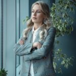 woman in gray blazer standing near green plant with a serious look