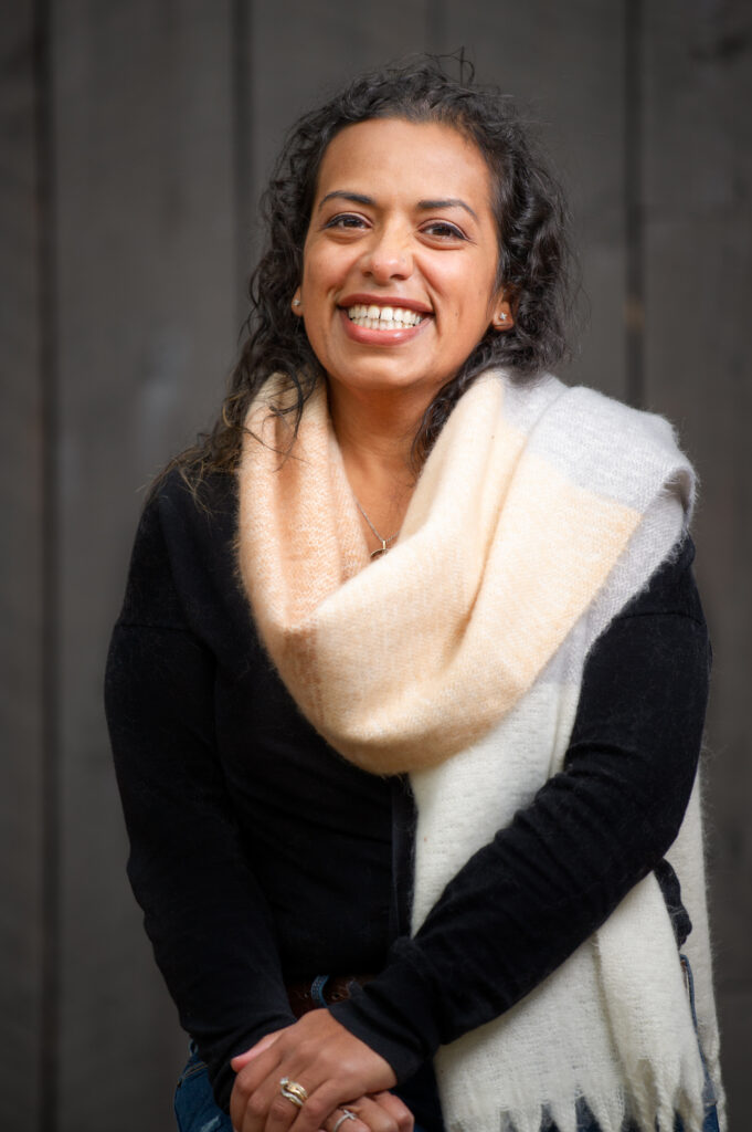 Julie Whitley smiles warmly in a cozy scarf and black sweater, standing against a rustic wooden background, radiating confidence and kindness.