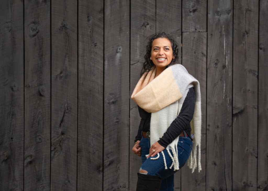 Julie Whitley smiling confidently in front of a rustic wooden wall, wearing a cozy color-block scarf and casual outfit, embodying warmth and approachability.