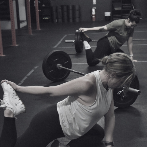 Two women stretch after a barbell workout in a gym, focusing on mobility and recovery in a strength training session.